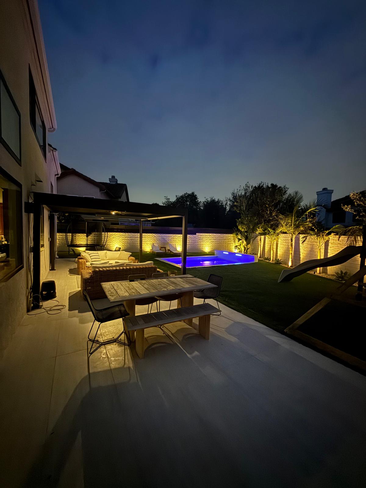 Backyard lit at dusk with blue pool and uplit landscaping