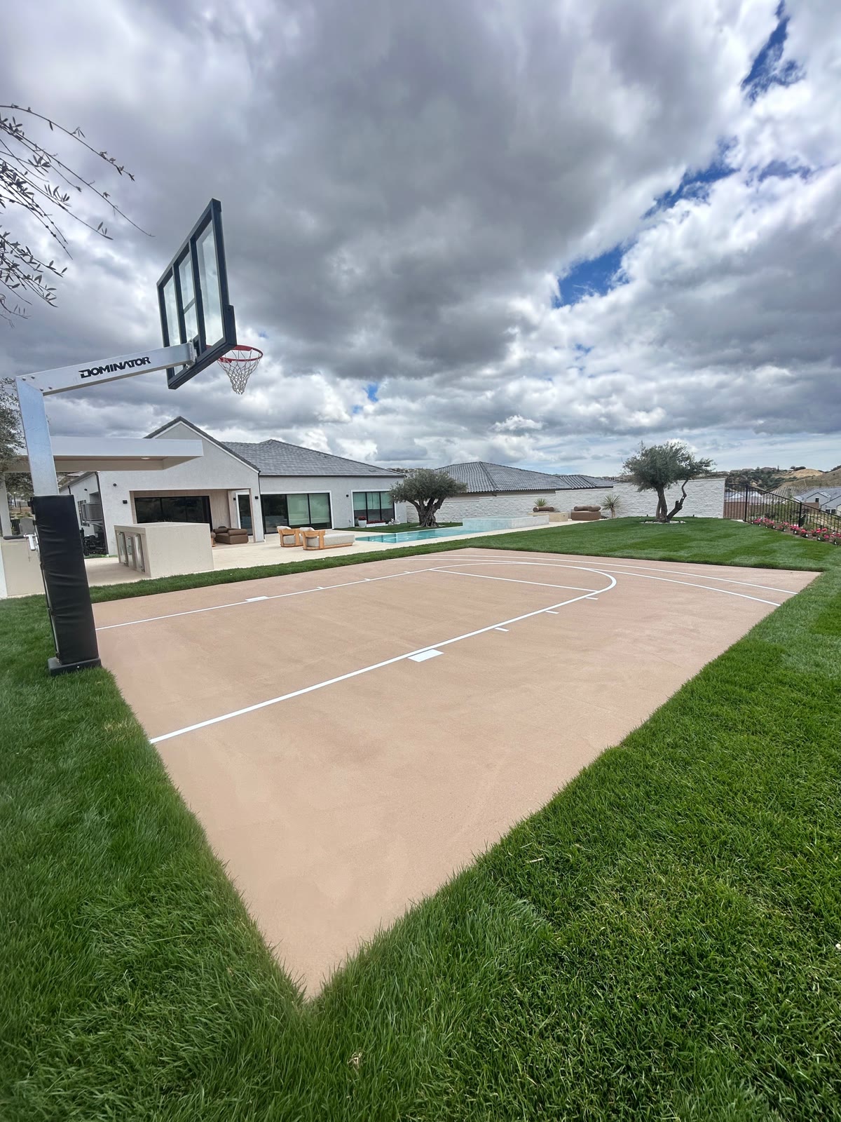 Tan backyard court with Dominator hoop, pool, and house beyond